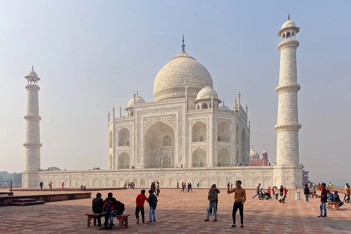 The Taj Mahal seen from the side across its marble plinth, with two minarets and visitors resting on benches in the foreground